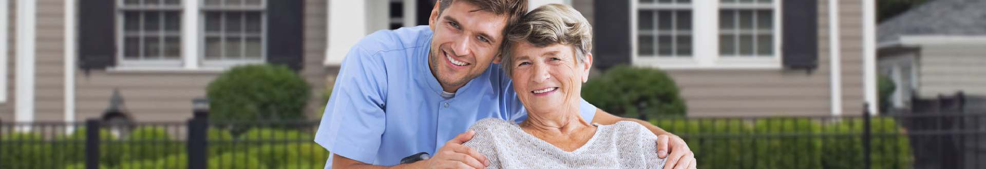 male caregiver and elder woman are smiling in front of a charming house